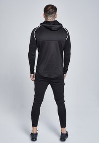 Schwarzer sportlicher Hoodie mit weißen Akzenten und langen Ärmeln, kombiniert mit schwarzen Jogginghosen mit schmal zulaufenden Beinen und Reißverschlussöffnungen an den Knöcheln.