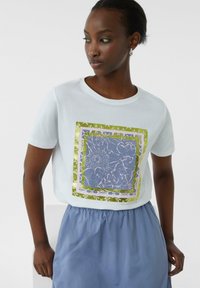 Witte T-shirt met kleurrijke bloemenprint omlijst in groen en paars. Gecombineerd met een lichtblauwe rok. Katoenen materiaal, korte mouwen.