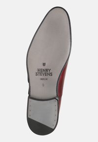 Sola della scarpa che mostra un materiale grigio liscio con un accento in pelle rossa. Il testo inciso reca la scritta "HENRY STEVENS BERLIN" con un numero di taglia.