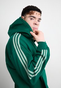 Hoodie verde com um tecido texturizado, apresentando três riscas verticais brancas em cada manga. Design com capuz e corte descontraído.
