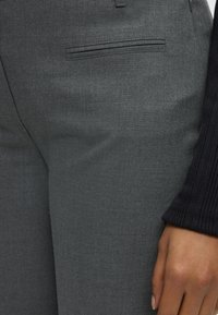 Gros plan d’un pantalon gris ajusté avec poche passepoilée et une main posée à côté portant un vêtement à manches côtelées noires.