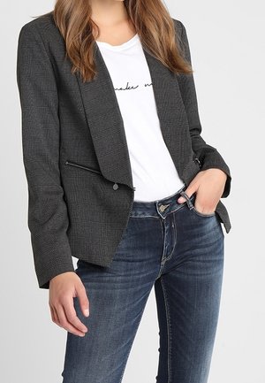 Donna che indossa un blazer a quadri grigio scuro sopra una t-shirt bianca con grafica e jeans blu scuro, con una mano nella tasca dei jeans.