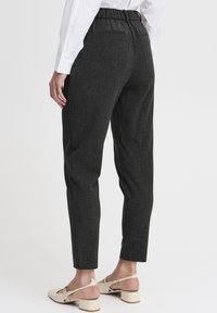 Donkergrijze op maat gemaakte broek van een gestructureerde stof, met een elastische tailleband, zijzakken en taps toelopende pijpen. G gedragen met lichtgekleurde schoenen.