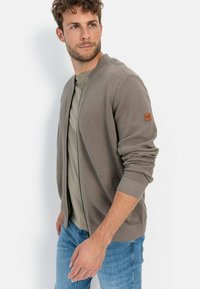 Lichtbruine gebreide ritscardigan met ribgebreide boorden en zoom, gecombineerd met een aansluitend olijfgroen t-shirt, gedragen met blauwe jeans.