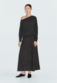 Übergroßer dunkelgrauer Strickpullover mit asymmetrischem Ausschnitt, kombiniert mit einem passenden Maxirock. Die Schuhe sind schwarze Slipper.