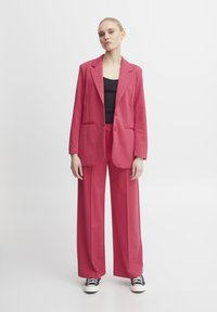 Rosa Blazer mit zwei frontalen Taschen, kombiniert mit passenden, weit geschnittenen Hosen. Darunter befindet sich ein schwarzes, tailliertes Oberteil. Das Outfit hat einen glatten Stoff und ein strukturiertes Design.