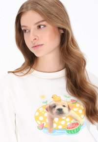 Fehér pulóver élénk nyomattal, amelyen egy kutya úszógumiban látható, sárga pöttyös mintával, kiskacsával és gyümölcsös díszítésekkel.