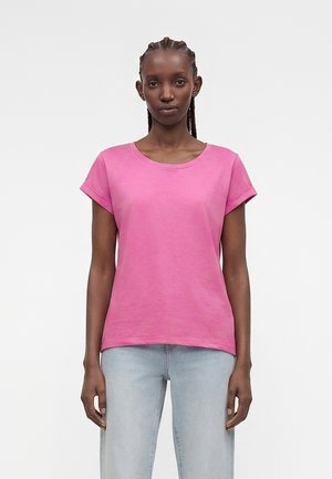 Femme aux cheveux tressés portant un t-shirt rose à manches courtes et un jean bleu clair, debout devant un fond blanc uni.