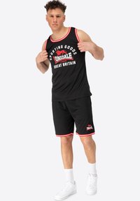 Čierny športový top na ramienka a šortky s červenými akcentmi. Obsahuje odvážny grafický potlač a logo. Model má obuté biele tenisky.