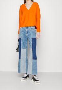 Oranger V-Ausschnitt-Pullover mit lockerer Passform; blaue Jeans mit unterschiedlichen Farbtönen und Patchwork-Design; schwarz-weiße Sneakers.