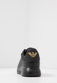 Black sneaker with a smooth leather finish, featuring a prominent gold logo on the heel and a ridged black sole for traction.