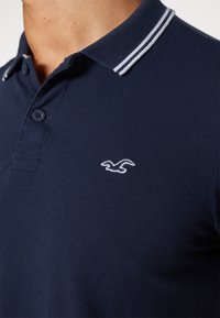 Námořnické polo tričko vyrobené z texturované tkaniny, s dvoubotkovou légou, kontrastními bílými pruhy na límci a bílým logem jako akcent.