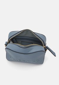 Sac en cuir bleu avec fermeture éclair dorée, présentant un logo en relief à l'avant et un intérieur doublé avec des poches pour l'organisation.