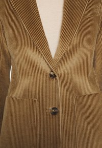 Brun fløjl blazer med klapkrave, to frontlommer og to knapper. Teksturen er ribbet med en blød overflade.