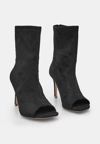 Juodi zomšiniai kulkšninių batai su atvirais pirštais, stiletto kulnais ir šoninėmis užtrauktukais, pateikti prieš vienodą šviesią fono.