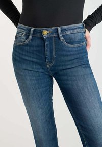 Mujer vestida con jeans de mezclilla azul de cintura alta y un botón dorado, combinados con una camiseta negra ajustada de manga larga, con las manos en los bolsillos.