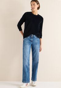 Musta samettinen pitkähihaisen neule, yhdistetty vaaleansinisin leveälahkeisiin farkkuihin. Valkoiset tennarit punaisilla yksityiskohdilla. Yksinkertainen tausta.