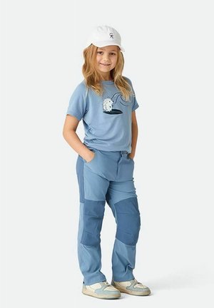 Barn stående med hænderne i lommerne iført lyseblå patchwork-bukser, blå t-shirt med pindsvinegrafik, hvid kasket og sneakers.