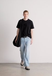 Melna kokvilnas T-krekls ar logo, gaiši zili džinsu bikses, pelēki apavi. Modelis nēsā melnu pleca somu un stāv neitrālā fonā.