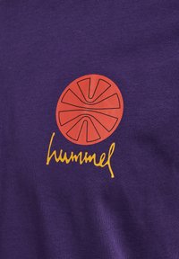 Fioletowa koszulka bawełniana z czerwonym okrągłym wzorem z czarnymi liniami i słowem "hummel" napisanym żółtą kursywą poniżej wzoru.