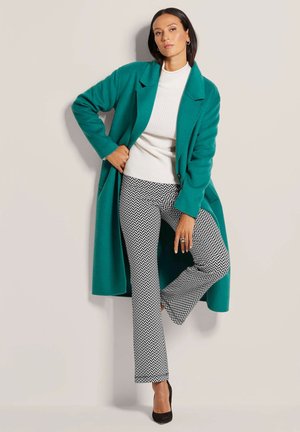Teal long coat over a white textured knit sweater, paired with black-and-white zigzag patterned pants and black high-heeled shoes.
