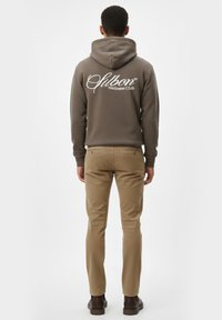 Sudadera marrón con logo blanco "Wellness Club" en la parte posterior, combinada con pantalones khaki beige y zapatos marrones. Tejido suave y casual.