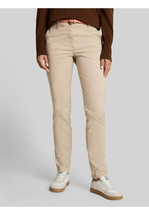 Personne portant un pantalon slim beige, un pull marron, une ceinture rose et des baskets blanches, debout devant un fond gris uni.