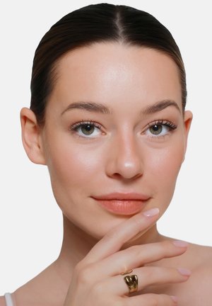 Giovane donna con pelle liscia, trucco leggero e occhi verdi, che tocca delicatamente il mento con la mano che indossa un anello d'oro largo.