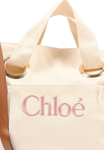 Beżowa torba typu tote z materiału płóciennego z krótkimi uchwytami i złotymi metalowymi pierścieniami, ozdobiona różowym haftowanym logo "Chloé" oraz widocznym brązowym skórzanym paskiem.