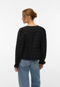 Femme aux cheveux roux relevés en chignon, portant un chemisier noir texturé à manches longues et un jean bleu clair, vue de dos sur un fond uni.