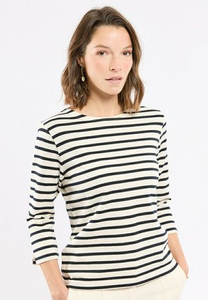 Femme aux cheveux bruns mi-longs portant un chemisier blanc à manches longues avec des rayures horizontales noires, mains dans les poches, sur fond blanc.