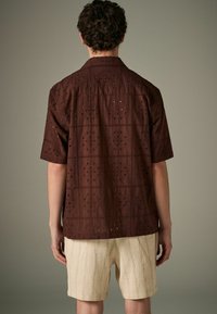 Chemise à manches courtes en œillets marron avec des motifs floraux carrés, dotée d'un col. Associée à un short crème léger et texturé.