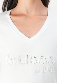 Biała bawełniana koszulka z dekoltem w kształcie litery V, ozdobiona wypukłym logo "Guess" z metalowymi ćwiekami oraz haftowanym detalem "est. 1981", o gładkiej fakturze.