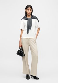 Γυναίκα που φοράει λευκή μπλούζα με κοντά μανίκια, μπεζ παντελόνι φαρδιάς γραμμής, μαύρα ψηλοτάκουνα παπούτσια, γκρι πουλόβερ πάνω στους ώμους και κρατά μια μικρή μαύρη τσάντα.