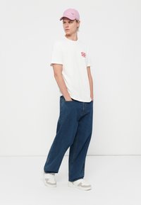 White cotton t-shirt with red graphic, blue wide-leg jeans, and pink cap. Grey and white sneakers complete the outfit.