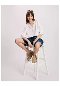 Biała koszula z długim rękawem typu henley, dżinsowe szorty i beżowe buty wsuwane. Model siedzi na białym stołku, nosząc jasne okulary przeciwsłoneczne.