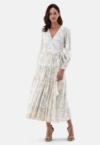 Weißes Maxikleid mit gelben und grauen Mustern, langen Ärmeln, tailliertem Bund und gestuftem Rock. Kombiniert mit grünen Riemchenheels.