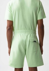 Jumpsuit verde de algodón de manga corta con cintura elástica, que cuenta con un bolsillo de parche en la parte posterior. Textura suave y corte relajado.