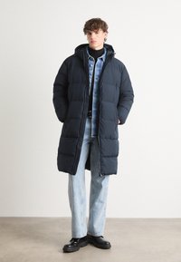 Long navy puffer coat with a hood, featuring a zippered front and large rectangular quilted design. Paired with a denim shirt and light jeans.