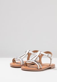 Sandalias blancas con acentos plateados, con un diseño de tiras y punta abierta. Fabricadas con suelas planas y una plantilla marrón. Con correa en el tobillo ajustable.