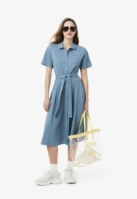 Vestito blu con bottoni, maniche corte e cintura in vita, abbinato a sneakers bianche, calzini bianchi e una borsa trasparente gialla con logo.