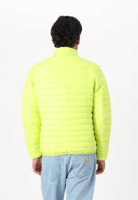 Neon yellow padded jacket with a high collar and horizontal stitching. Worn over light blue jeans, showcasing a simple design.
