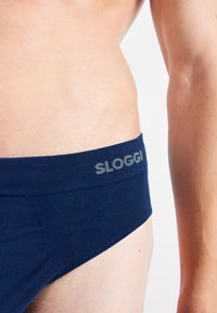 Nahaufnahme einer Person, die navyblaue Unterwäsche mit dem "SLOGGI"-Logo am Bund vor einem weißen Hintergrund trägt.