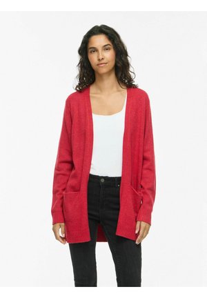 Femme aux longs cheveux bouclés et foncés portant un cardigan rouge ouvert, un haut blanc et un jean noir taille haute, debout sur un fond blanc uni.