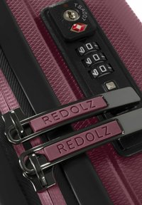 Dettaglio delle cerniere di una valigia color bordeaux con il marchio REDOLZ, bloccate da un lucchetto combinato TSA nero impostato su 000.