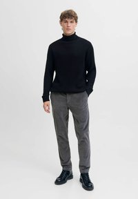 Schwarzer gerippter Rollkragenpullover, graue Cordhose und schwarze Schnürstiefel, vor einem schlichten weißen Hintergrund stehend.