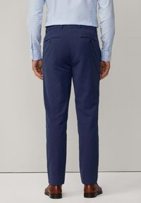 Pantaloni navy con una texture liscia, dotati di due tasche posteriori con bottoni e una vestibilità sartoriale, abbinati a scarpe in pelle marrone.