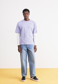 Lilla kortærmet t-shirt lagd over en hvid langærmet skjorte, parret med lyseblå jeans og sorte sneakers, stående på et gult gulv.