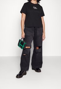 Camiseta negra corta con el logo de "Tommy Jeans", jeans anchos negros desgastados con rasgaduras en las rodillas, y un pequeño bolso verde y negro. Botas negras.