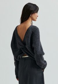 Žena na sobě má tmavě šedý pletený svetr s volnými zády a černé kalhoty s vysokým pasem, stojící proti jednobarevnému pozadí.